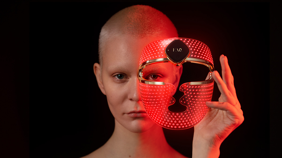 a fit young woman, no makeup, blond buzz cut, holding the FAQ 202 led face mask turned on and glowing red, one eye  is looking through the mask eye slit